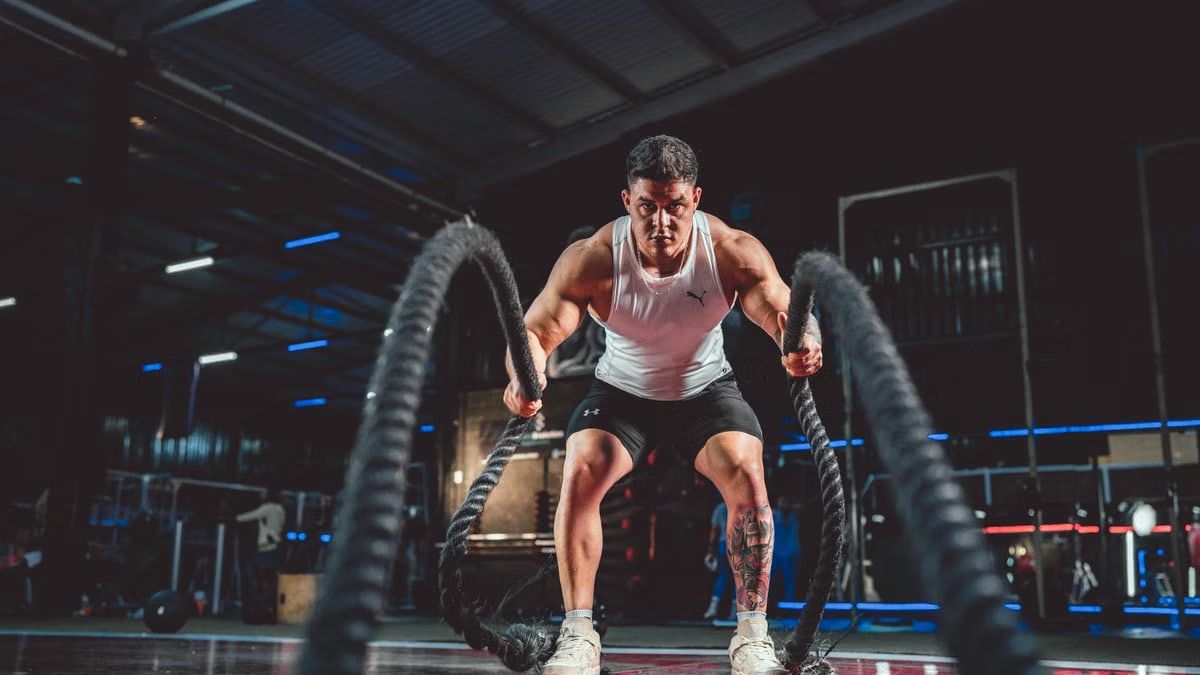 Focused man in white tank top performing battle rope exercise in a modern gym.