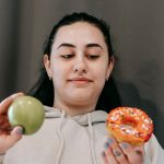 Content young female in hoodie deciding between sweet unhealthy doughnut and green ripe apple in light room