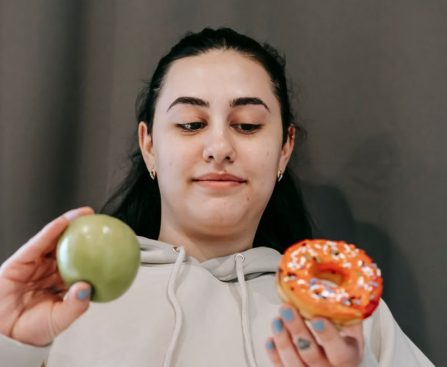 Content young female in hoodie deciding between sweet unhealthy doughnut and green ripe apple in light room